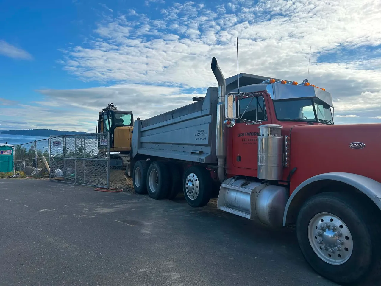 A Gritworks Trucking dump truck, company name lettered on the door, at a waterfront job site.